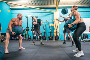 four athletes one male lifting a heavy tfench sandball, one adaptive athlete holding a sandball, one female athlete lifting a sandball and one male athlete tossing a sandball 